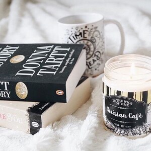 Puede incluir: Una vela encendida en un tarro de cristal con una etiqueta negra y dorada que dice "Parisian Cafe" est&aacute; junto a dos libros y una taza. Los libros se titulan "The Secret History" y "Me Before You". La taza tiene las palabras "Time Read".