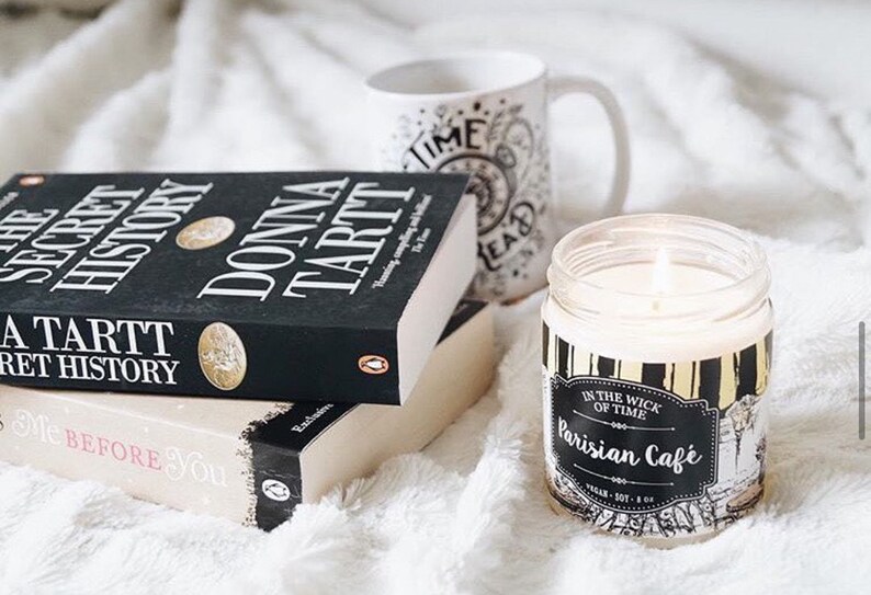 Puede incluir: Una vela encendida en un tarro de cristal con una etiqueta negra y dorada que dice "Parisian Cafe" est&aacute; junto a dos libros y una taza. Los libros se titulan "The Secret History" y "Me Before You". La taza tiene las palabras "Time Read".