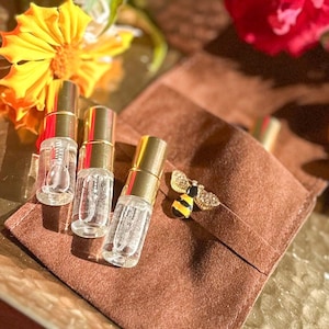 May include: Three small, clear perfume bottles with gold caps are arranged on a brown suede pouch. A gold and enamel bee pin rests on the pouch. Bright yellow and red flowers are in the background.