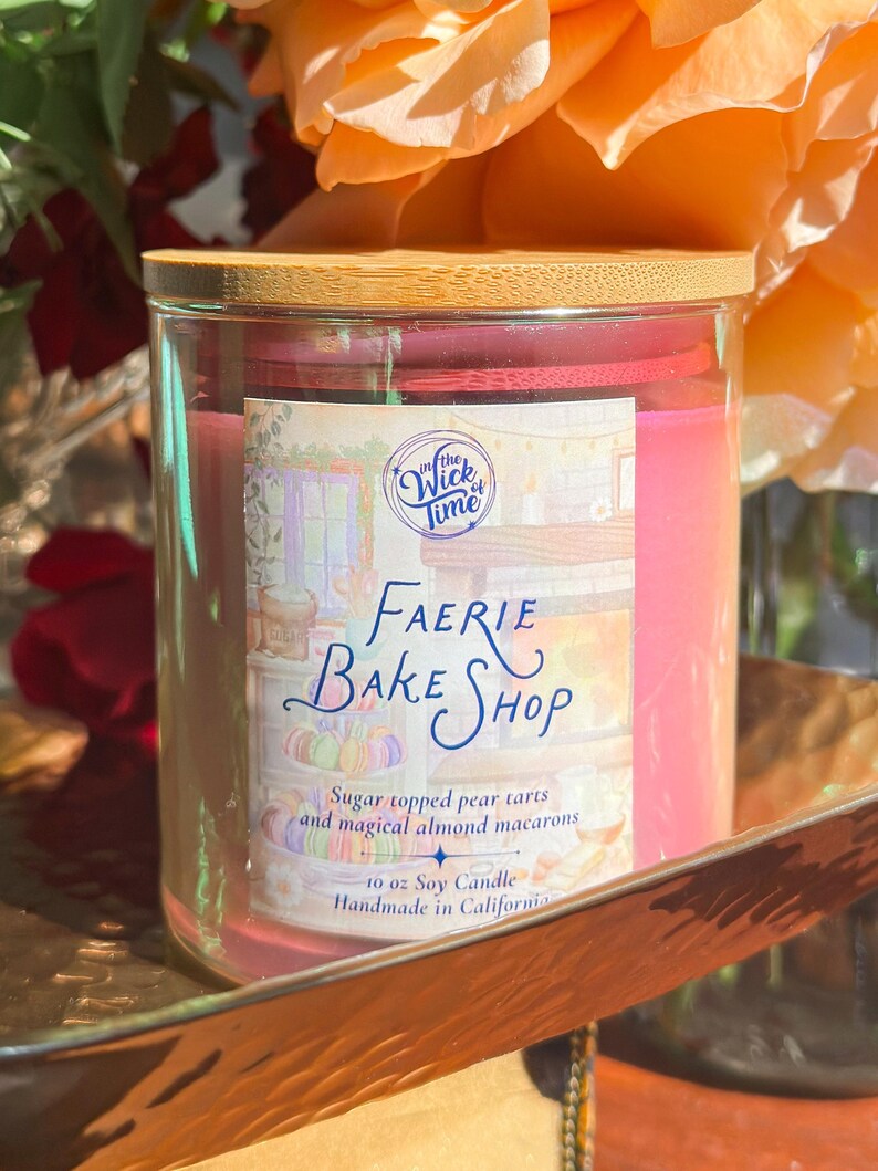 May include: A glass candle jar with a wooden lid. The label reads "Faerie Bake Shop" with text about pear tarts and almond macarons. The candle is pink and is 10 oz. Handmade in California.