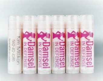May include: Six clear tubes of lip balm with pink labels that say "Damsel in Defense" and a pink silhouette of a woman. The label on the first tube says "Kim Millikan 562-412-4285 kimmillikan.com".
