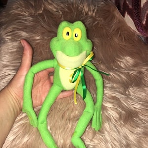 Puede incluir: Juguete de peluche de rana verde con grandes ojos amarillos y una cinta amarilla y verde alrededor del cuello.