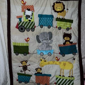 May include: A beige baby quilt featuring a train design with various animals in the train cars. The animals include a zebra, lion, elephant, bear, monkey, and giraffe. The quilt has a brown border and is decorated with stars and birds.