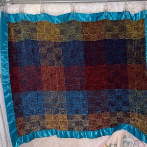 May include: A colorful patchwork quilt with a teal satin border. The quilt features squares of red, blue, and gold, creating a geometric pattern. The quilt is hanging from a curtain rod, showcasing its design and texture.