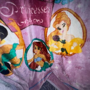 K&ouml;nnte beinhalten: Eine rosa Fleece-Decke mit Prinzessinnen-Motiv. Die Decke zeigt drei Prinzessinnen in ovalen Rahmen. Die Prinzessinnen sind Jasmin, Belle und Arielle. Der Text "Princesses Only" befindet sich oben auf der Decke.