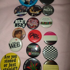 May include: A collection of 15 round button pins featuring various band logos and slogans. The pins are colorful and feature a variety of designs, including the Ramones, The Police, Sex Pistols, and The Cars.