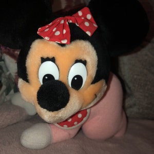 May include: A plush toy of Minnie Mouse wearing a pink and white polka dot onesie and a red and white polka dot bow. The toy has large black ears and a black nose.