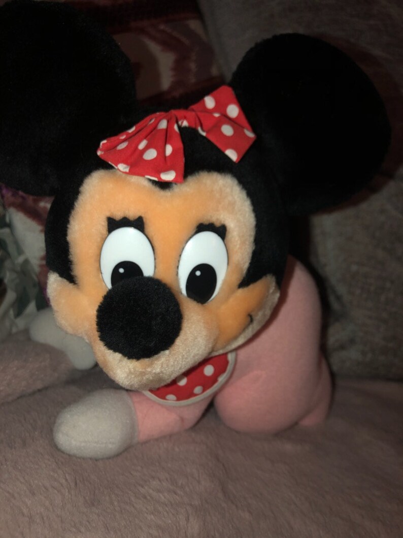 May include: A plush toy of Minnie Mouse wearing a pink and white polka dot onesie and a red and white polka dot bow. The toy has large black ears and a black nose.