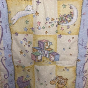 May include: A baby quilt with a light blue border and a yellow and white checkered pattern. The quilt features a lamb, a crescent moon, stars, and blocks that spell out "LOVE". The text "Welcome to the world" is written on the top border.