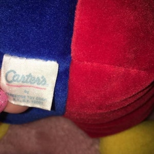 Op de afbeelding: Een rood en blauw pluche speelgoed met een geel gezicht. Het speelgoed heeft een label met de tekst "Carter's" erop.