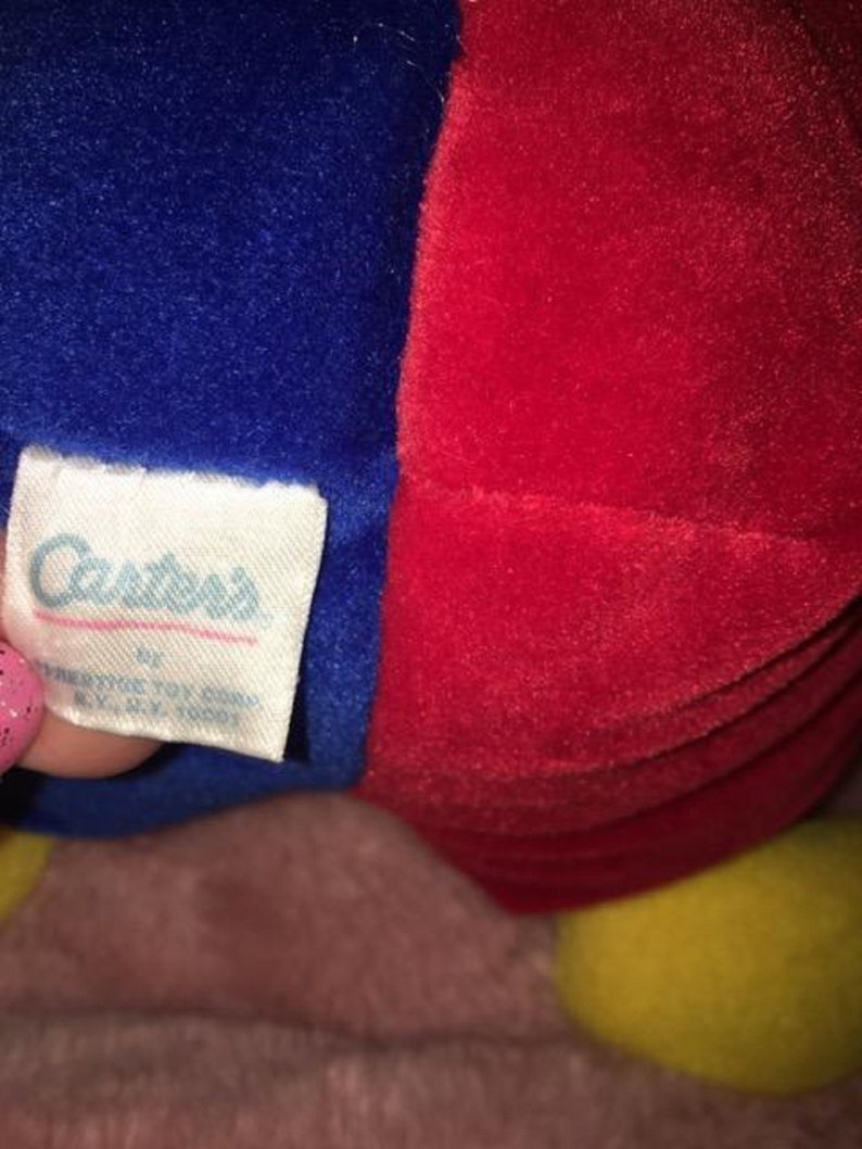 Op de afbeelding: Een rood en blauw pluche speelgoed met een geel gezicht. Het speelgoed heeft een label met de tekst "Carter's" erop.