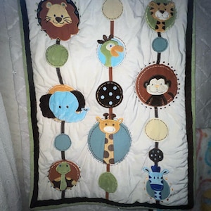 May include: A white baby quilt with a brown border and green trim. The quilt features a variety of animal appliques, including a lion, giraffe, monkey, elephant, turtle, and zebra. The animals are arranged in a circular pattern on the quilt.