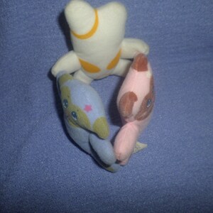 May include: Three plush toys in a circle. The toy on the left is blue and green with a pink star on its chest. The toy on the right is pink with brown ears and a brown face. The toy on the top is white with a yellow crescent moon on its chest.