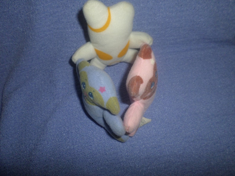 May include: Three plush toys in a circle. The toy on the left is blue and green with a pink star on its chest. The toy on the right is pink with brown ears and a brown face. The toy on the top is white with a yellow crescent moon on its chest.