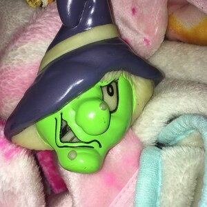 May include: A green witch figurine with a purple hat and a mischievous expression. The figurine is made of plastic and has a light green glow in the dark feature.