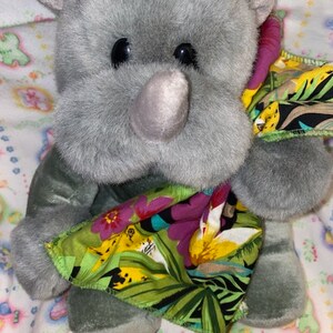 May include: A gray plush rhinoceros toy with a black nose and large black eyes. The rhino is wearing a green, yellow, and pink patterned fabric blanket.