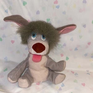 Puede incluir: Un juguete de peluche gris y marrón en forma de perro con la lengua rosa que sobresale. El juguete tiene una cabeza peluda y grandes ojos azules.