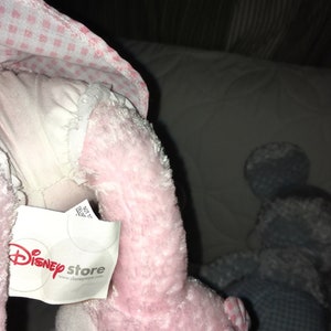 Op de afbeelding: Roze en wit gingham stof met een roze en wit pluche konijn speelgoed. Het konijn heeft een witte strik op zijn oor en een wit label met de tekst "Disney store" met het website adres "www.disneystore.com".