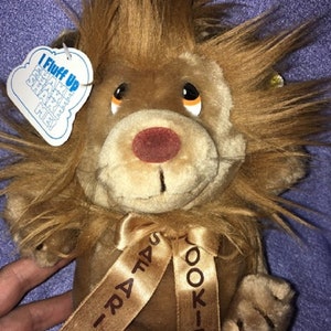 May include: A brown plush lion with a brown bow tied around its neck. The bow has the words "I-RAT-A-TUS" and "KOOKIE" printed on it. The lion has a tag that says "I Fluff Up Shake Shake Me!"