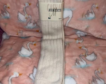May include: A pair of cream-colored ribbed socks with a band that reads "Casual Moments". The socks are folded and placed on a pink blanket with a swan pattern.