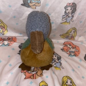May include: A plush toy shaped like a bird with a brown body, gray head, and green wings. The toy is sitting on a white blanket with a pattern of stars and Disney princesses.