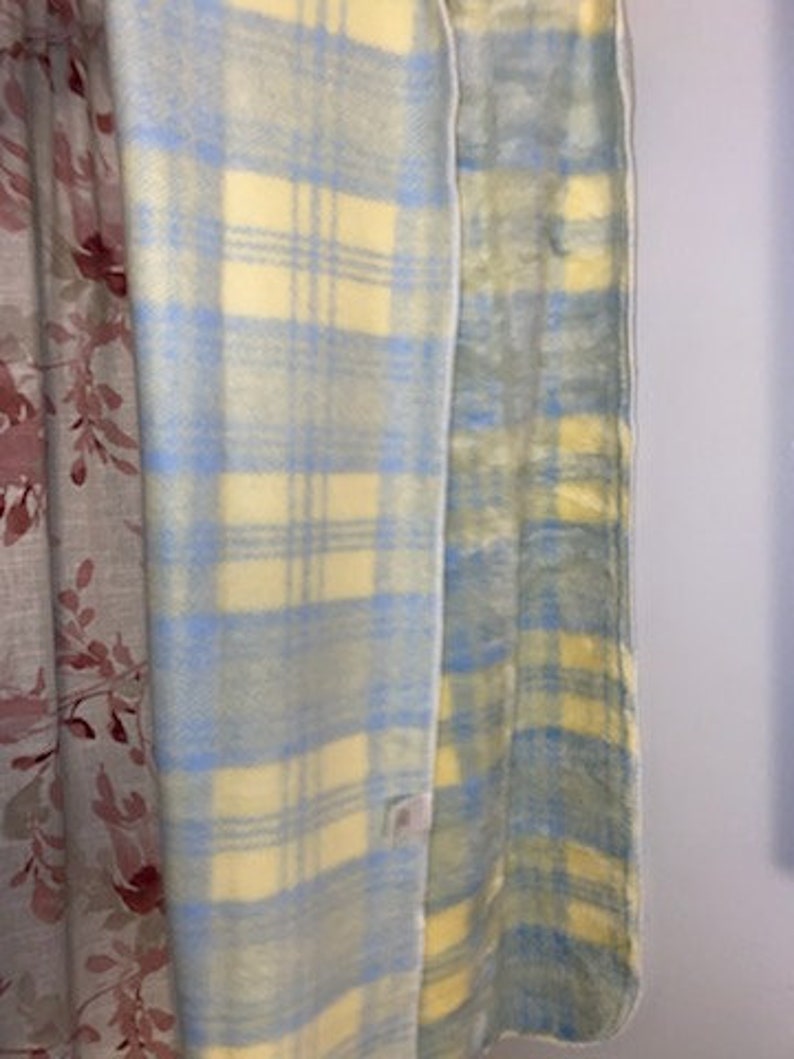 May include: A blue and yellow plaid blanket with a white border. The blanket is folded in half and is hanging on a wall.
