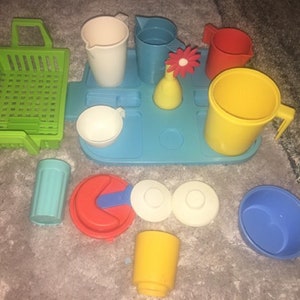 May include: A set of colorful plastic toy dishes and cups, including a green basket, a blue tray, a yellow pitcher, a red cup, a white cup, a blue cup, a white bowl, a blue bowl, and a yellow bowl.