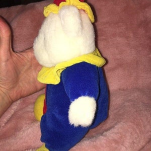 Op de afbeelding: Een witte pluche clown met een rode en gele hoed, een blauwe broek en gele schoenen. De clown draagt een gele kraag en heeft een wit gezicht.