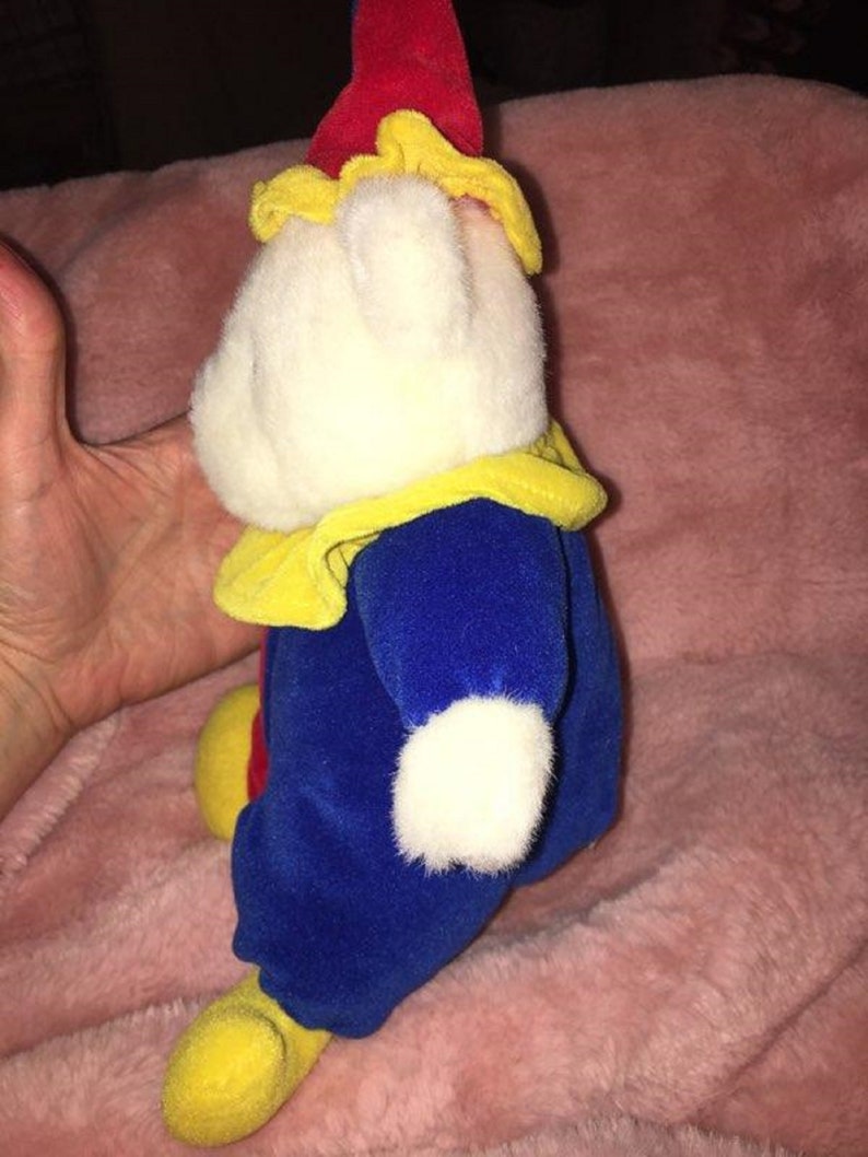 Op de afbeelding: Een witte pluche clown met een rode en gele hoed, een blauwe broek en gele schoenen. De clown draagt een gele kraag en heeft een wit gezicht.
