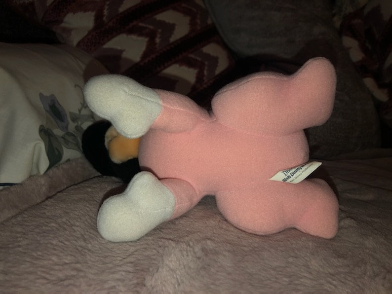 May include: Pink plush toy with white feet and a Disney Store tag. The toy is lying on its back with its legs in the air.