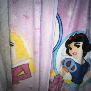 K&ouml;nnte beinhalten: Eine rosa Fleece-Decke mit einem violetten Rand, die Schneewittchen und Dornr&ouml;schen zeigt. Die Decke hat ein Muster aus wei&szlig;en Sternen.