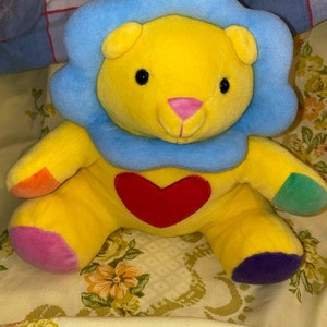 May include: A yellow plush lion with a blue mane and a red heart on its chest. The lion has colorful patches on its arms and legs.