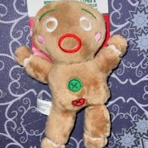 May include: A plush gingerbread man toy with a smiling face, rosy cheeks, and a red mouth. It has a green button and a red cross on its body. The toy is attached to a white and red tag that reads "Pull-String Gingerbread Man".
