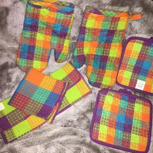 May include: A collection of kitchen textiles, comprising two oven gloves, two pot holders, and two matching tea towels. The items display a bright plaid design in purple, orange, blue, and green. The pot holders have a purple trim.