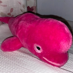 May include: A plush pink whale toy with a white belly and black eyes. The whale is lying on a white surface.
