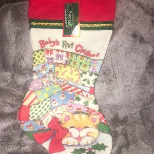 May include: A Christmas stocking with a red cuff and a white body. The stocking is decorated with a patchwork design and the words "Baby's First Christmas." A cartoon kitten with a red bow is featured on the toe.