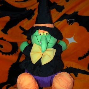 May include: A green witch plush toy with a black hat, yellow bow, and orange feet. The witch is sitting on an orange and black blanket with black cat silhouettes.