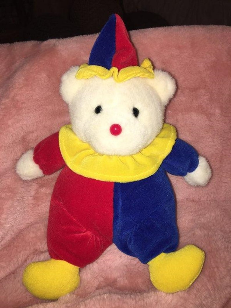 Op de afbeelding: Een witte pluche teddybeer verkleed als clown. De beer draagt een rood en blauw narrenkostuum met gele accenten. De beer heeft een rode neus en zwarte ogen.