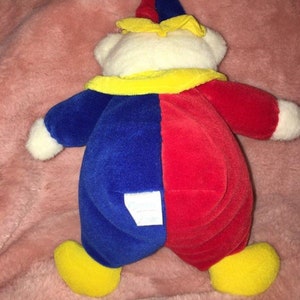 Op de afbeelding: Een pluche clownbeer met een rood en blauw kostuum, gele hoed en schoenen. De beer heeft een wit gezicht en een label op zijn rug.