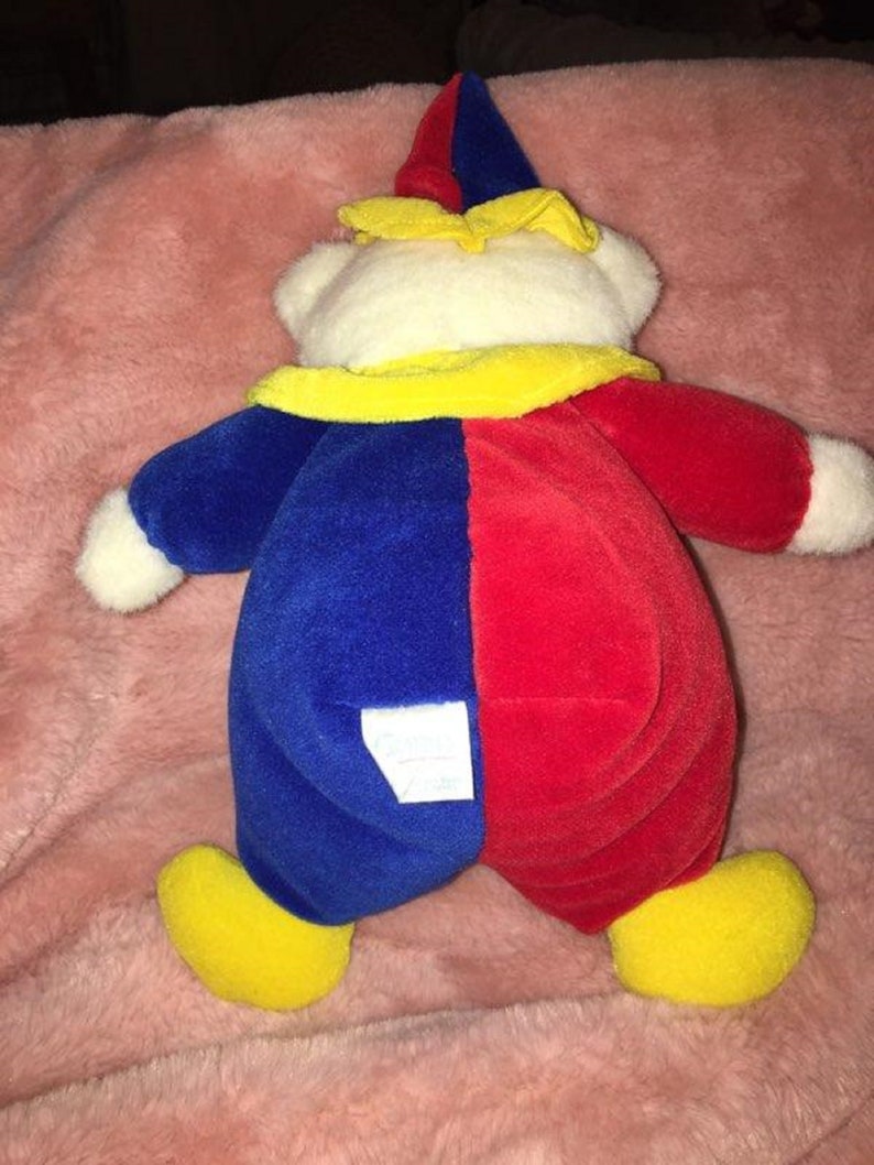 Op de afbeelding: Een pluche clownbeer met een rood en blauw kostuum, gele hoed en schoenen. De beer heeft een wit gezicht en een label op zijn rug.
