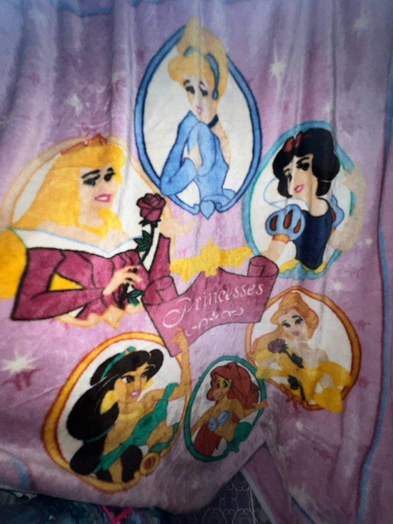 K&ouml;nnte beinhalten: Eine lilafarbene Fleece-Decke mit einem rosa, gelben und wei&szlig;en Design mit Disney-Prinzessinnen. Die Decke hat ein Banner mit der Aufschrift "Princesses of Dreams".