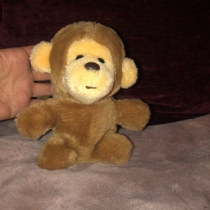 May include: A plush brown monkey toy with a light brown face, ears, and hands. The monkey has a dark brown nose and eyes. The toy is sitting on a pink, fuzzy surface.