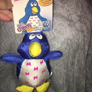 May include: A blue and white plush penguin toy with a yellow beak and feet, and pink bow accents. The toy is attached to a cardboard tag that reads "Penny the penguin" and "I'm FUN in the tub, pool and bedtime!"