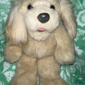 May include: A plush golden retriever puppy toy with brown eyes and a pink tongue sticking out. The toy is sitting on a green and white patterned blanket.