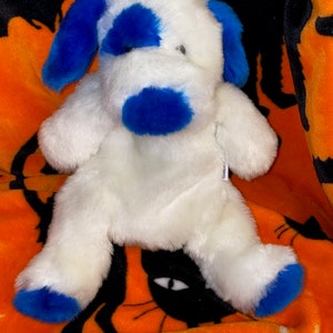 May include: White plush dog toy with blue ears, nose, and spots. The toy is sitting on an orange and black blanket.