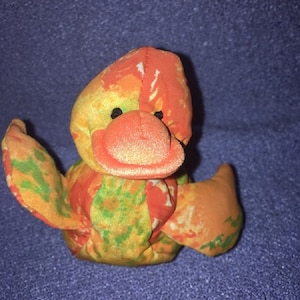 May include: A small, colourful plush duck toy with a bright orange beak and eyes. The duck is made of a patchwork fabric with orange, yellow, green, and white patterns.