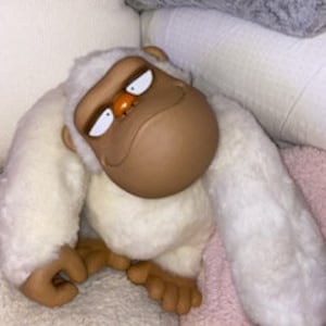 May include: A white plush monkey with brown fur on its face and hands. The monkey has a grumpy expression and is sitting on a white and pink blanket.