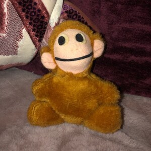 May include: A plush toy monkey with a light brown face, large black eyes, and a stitched black smile. The monkey has orange fur and small, rounded ears. It is sitting on a soft, light purple surface.