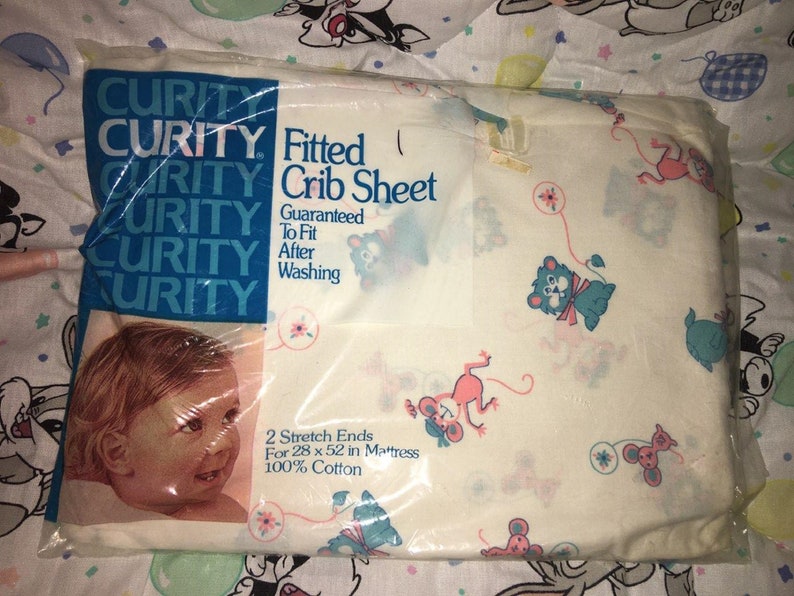 May include: A packaged Curity fitted crib sheet. The sheet is white with a pattern of cartoon animals in pink, blue, and green. The packaging includes the Curity logo and text that reads "Fitted Crib Sheet" and "Guaranteed To Fit After Washing."