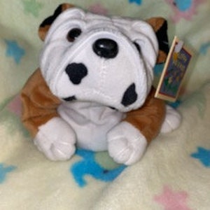 May include: A plush brown and white bulldog with black spots. The bulldog is sitting with its paws in front of it. The bulldog has a tag that says "The Original Plush Pals".
